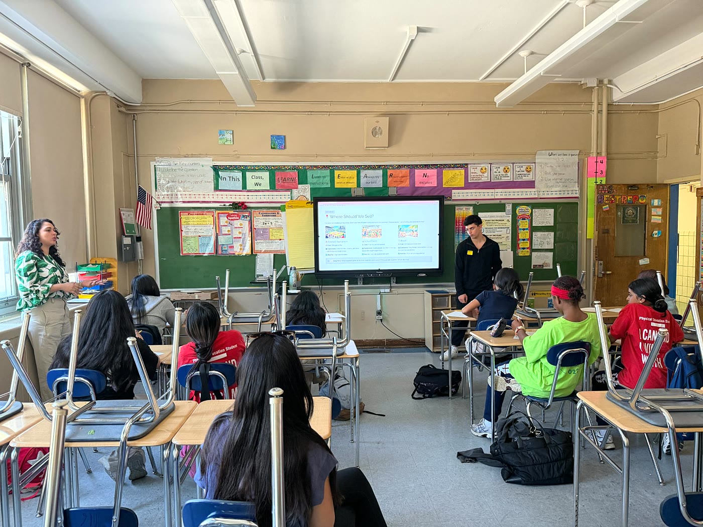 A teacher and a GUS presenter lead a classroom of students through a slide titled Where Should We Sell?
